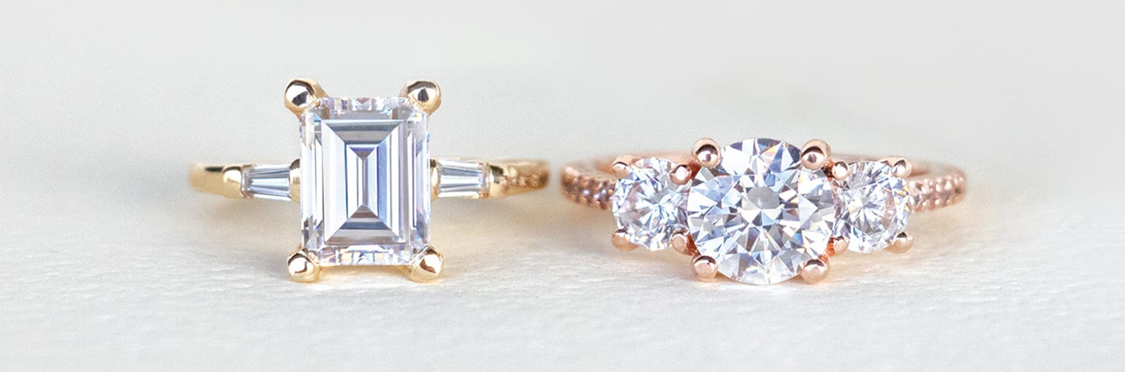 An emerald cut engagement ring in a three stone setting and a round brilliant cut engagement ring in a three stone setting